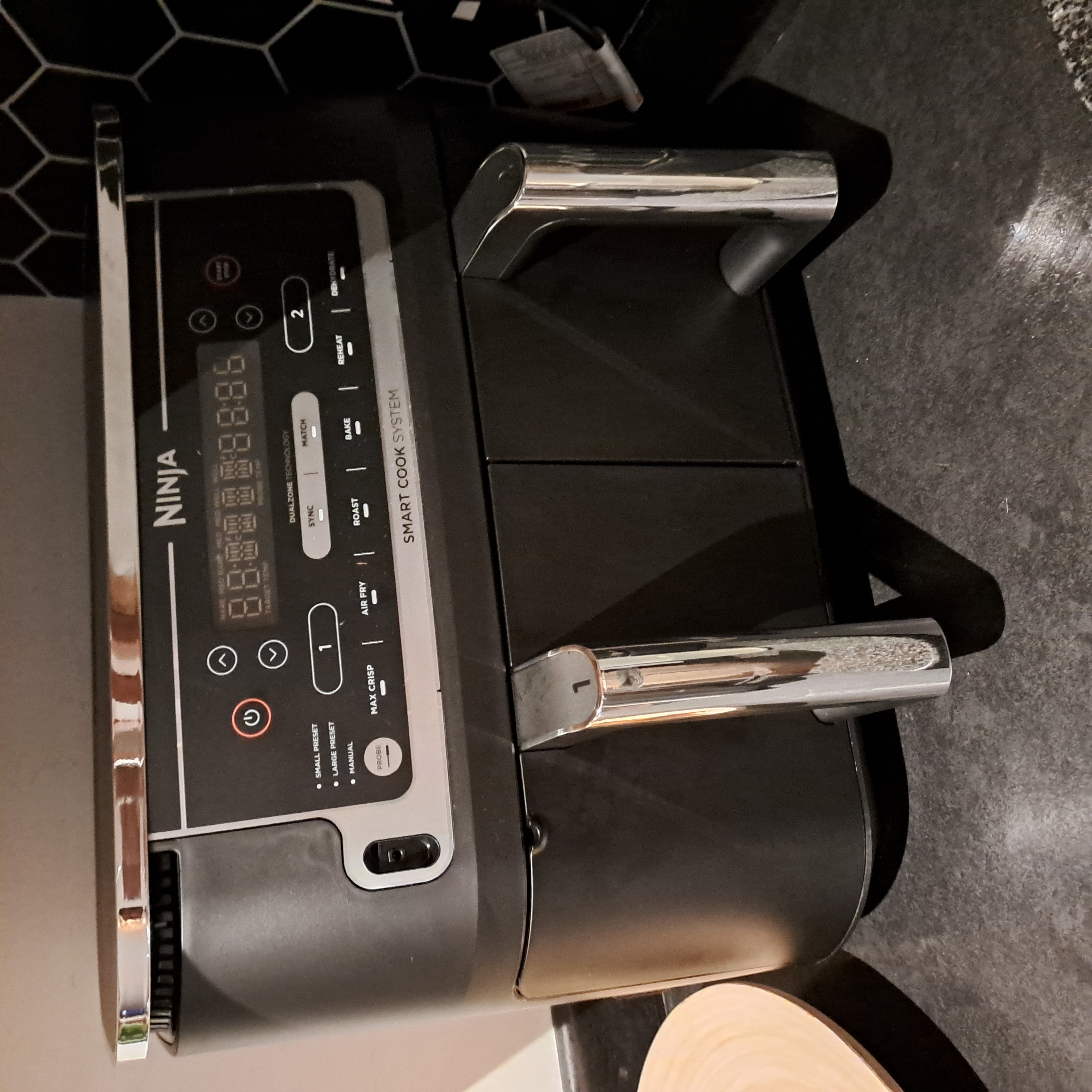 a square photo of a black airfryer of the ninja brand. it is sitting on a black textured counter top, with a white wall behind it and a wall of black hexagonal tiles beside it