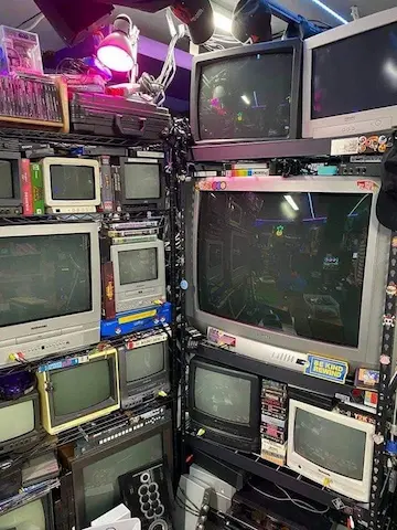 a photo of 2 shelves meeting at a corner, packed full of crts of different sizes, as well as cases of cassetts and disks and the likes. stickers decorate some of the crts and shelves